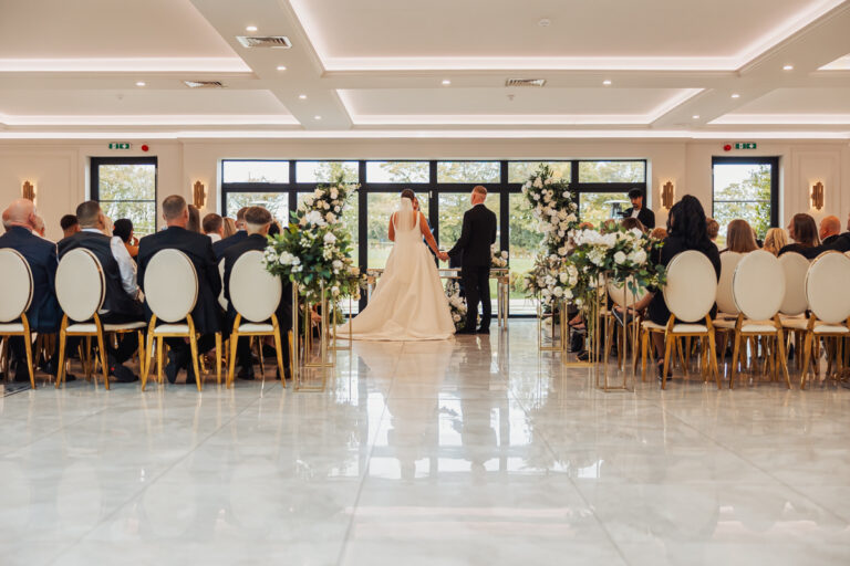 A bride in a white gown and a man in a dark suit stand at the front of a room, facing rows of seated guests during a wedding ceremony at Walton View Hotel, Bradford