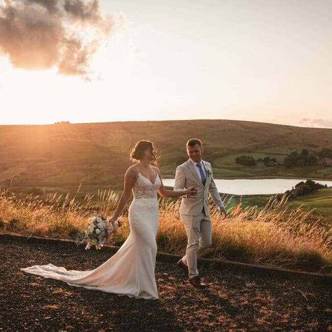a golden sunset photo taken at saddleworh hotel as a bride and groom are walking with castleshaw lower reservoir in the background | Yorkshire wedding Photographer