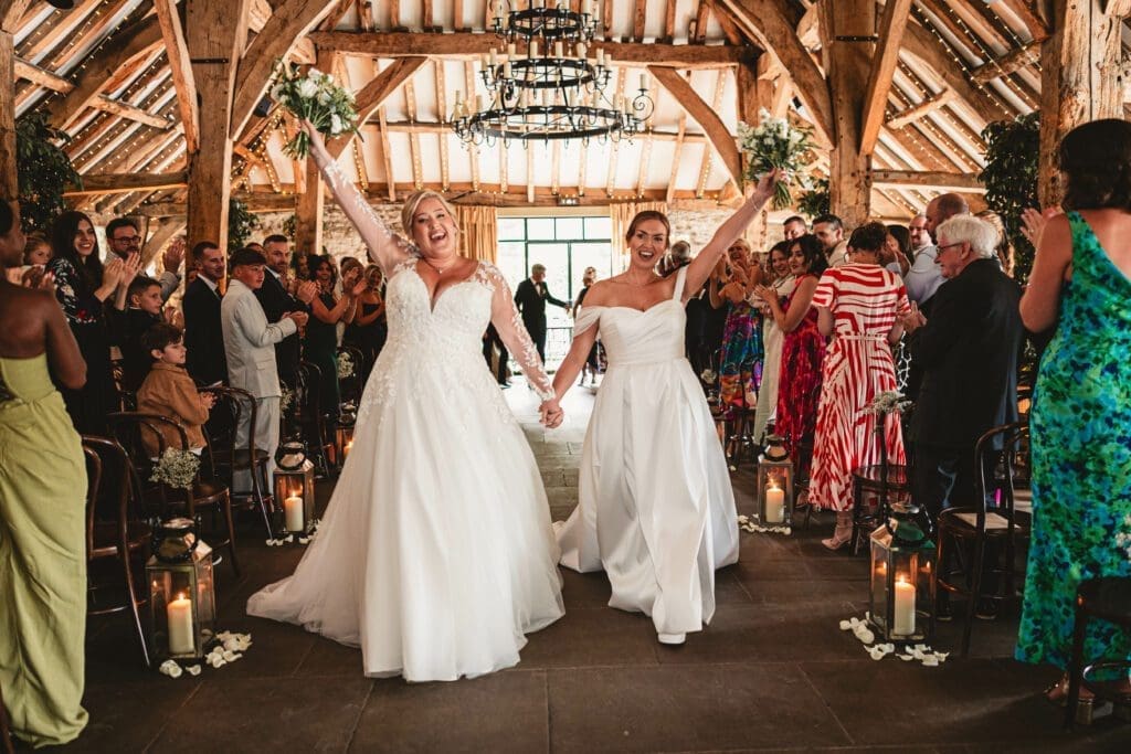 2 Brides walking out together , flowers in the air either side after they had got married at the tithe barn as there guests clap and share there joy for the big day