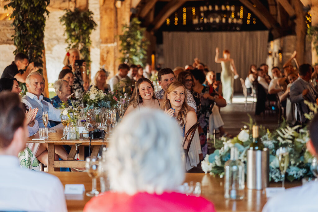 A lively north yorkshire wedding reception in a rustic hall, with guests seated at decorated tables, smiling and clapping while watching a speech. Warm lighting and greenery fill the Tithe Barn at Bolton Abbey, creating a festive atmosphere.