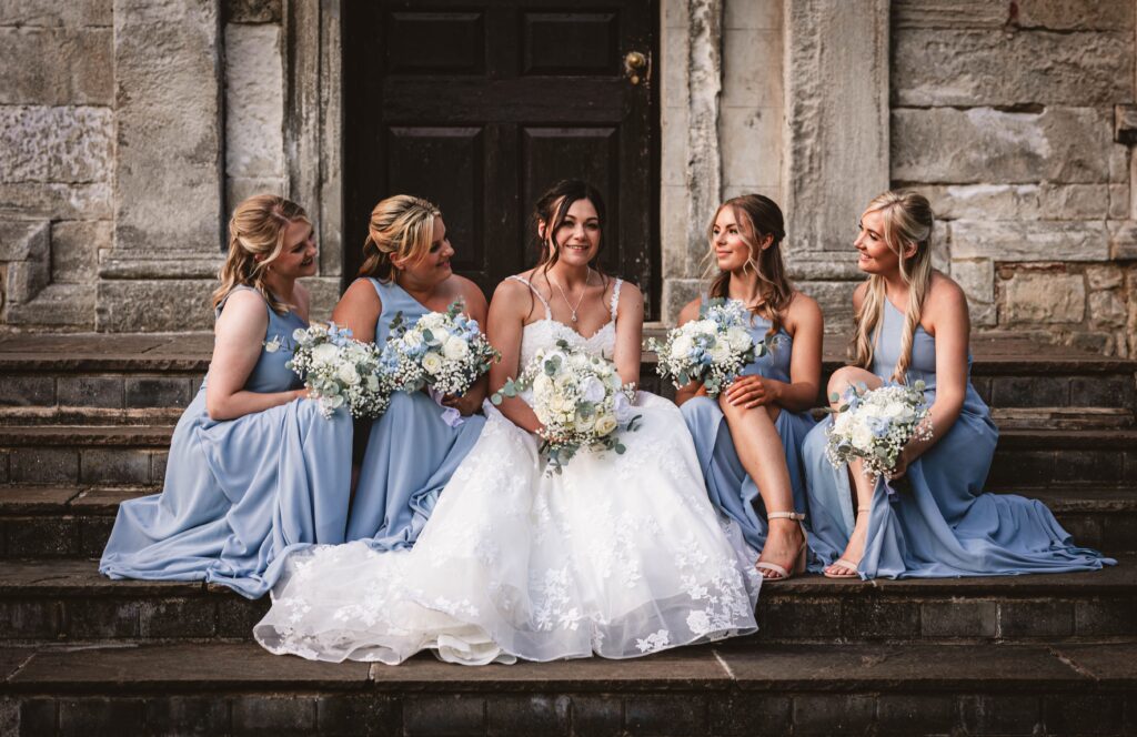 A bride in a white dress sits on stone steps with four bridesmaids in light blue dresses, all holding bouquets and smiling together in front of a stone building with a dark door.
