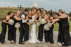 A joyful bride in a white dress holds a bouquet aloft, surrounded by bridesmaids and a groomsman in black, celebrating together and posing playfully outdoors on a stone patio at The Moorlands Halifax.