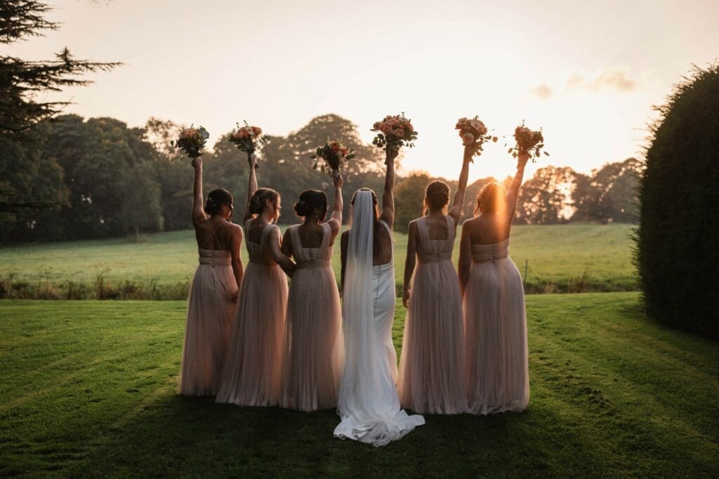 A bride in a white dress stands with five bridesmaids in pale pink dresses, all facing away and raising bouquets in the air, with a sunset and green field in the background.