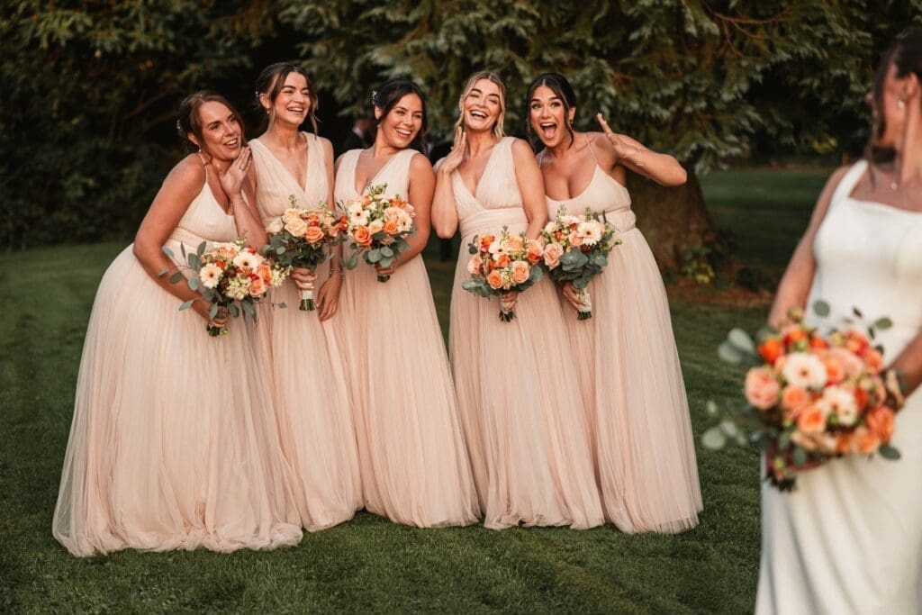 Five bridesmaids in light pink dresses stand smiling and holding bouquets, posing playfully outdoors on grass. A woman in a white dress, likely the bride, is partially visible holding flowers in the foreground.