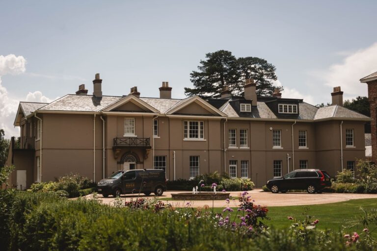 Burtonfields Hall is a large beige mansion with multiple chimneys and a well-kept garden in front. Two cars are parked on the driveway, while flowering plants line the circular entrance. Trees and a blue sky complete the picturesque setting.