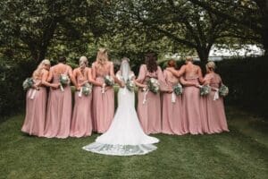 A bride in a white gown stands with her bridesmaids, all in matching blush pink dresses, backs to the camera, holding bouquets under leafy trees on a green lawn.