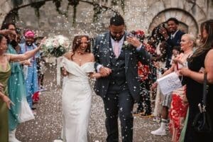 A joyful bride and groom walk arm in arm at Hazlewood Castle while guests toss white confetti. The bride holds a bouquet and laughs, both are dressed formally, surrounded by smiling friends and family outdoors.