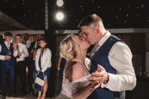 The newlyweds share a tender kiss on the dance floor at Moorlands Halifax, surrounded by smiling guests. The bride's off-shoulder dress and the groom's suit vest blend perfectly with the romantic setting. As the string lights twinkle above, some guests capture this wedding photography moment while others clap joyfully.