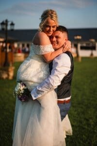 A joyful couple celebrates outdoors, captured in exquisite wedding photography. The groom, in a vest and shirt, holds the bride, who wears a lace gown and clutches a bouquet. They smile warmly at sunset on the picturesque Moorlands Halifax backdrop, with a blurred building and lamp post behind.