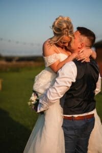 Captured in the enchanting scenery of Moorlands Halifax, a couple is lovingly embracing during their wedding. The groom, in a dark vest and white shirt, lifts the bride, who dazzles in her white dress and bouquet. This magical moment unfolds as the sun sets over the breathtaking moorlands.