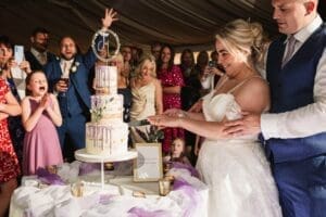 A bride and groom smile joyfully while cutting their wedding cake, expertly captured by a wedding photography team. The tiered cake, decorated with lavender sprigs and a topper with initials, stands in a festive tented venue resembling the serene beauty of Moorlands Halifax.
