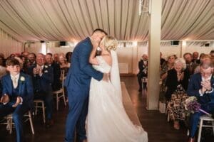 Amidst the enchanting setting of Moorlands Halifax, a bride and groom share a tender kiss during their wedding ceremony inside a tent. Surrounded by clapping guests, the moment is flawlessly captured through exquisite wedding photography as the groom dons a blue suit and the bride shines in her white gown with veil.