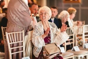 An elderly woman in a white lace cardigan sits in a wooden chair, holding a tan handbag, smiling and clapping. Around her, guests dressed in formal attire are seated in rows inside a brightly lit room captured perfectly by wedding photography at Moorlands Halifax.