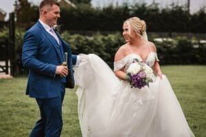 A bride and groom smile at each other while walking on the lush grass at Moorlands Halifax. The bride holds a bouquet of purple and white flowers, and the groom carries a drink in his left hand. Both are dressed formally, with trees and a fence enhancing this perfect moment of wedding photography bliss.