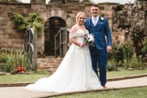 The bride and groom stand together outdoors in front of an old stone structure, captured beautifully in their wedding photography. The bride, holding a bouquet and dressed in a white gown, stands beside the groom in a blue suit. They smile amid the lush greenery of Moorlands Halifax.