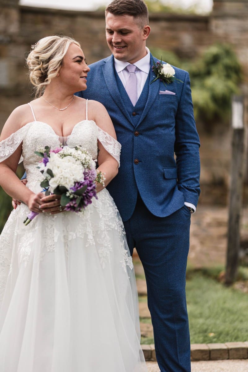A bride and groom stand outdoors in formal attire, set against the enchanting backdrop of the Moorlands Halifax. The bride wears a white gown and holds a bouquet of purple and white flowers. The groom is in a blue suit with a lavender tie. They gaze at each other, smiling as their moment is captured in exquisite wedding photography.