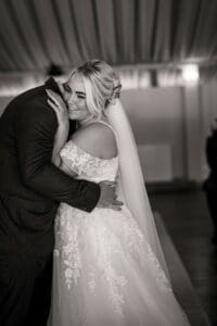 A black and white image of a bride in an off-the-shoulder lace gown, captured flawlessly by Moorlands Halifax wedding photography, embracing a groom in a suit. She smiles warmly, eyes closed, with a veil cascading down her back. The softly blurred background creates an intimate atmosphere.