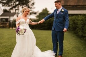 A bride and groom stand on a grassy field, captured perfectly in wedding photography. The bride, in a white dress, holds a bouquet of white and purple flowers, while the groom in a blue suit clasps her hand. Trees and the charming Moorlands Halifax are visible in the background.