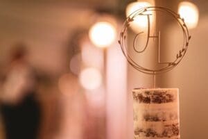 A single-tier, semi-naked cake stands elegantly, crowned with a monogram topper featuring the letters J and L inside a circular wreath design. Captured in the enchanting glow of wedding photography at Moorlands Halifax, the softly blurred background adds a touch of romance.