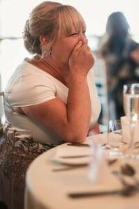 A woman with blonde hair, wearing a white dress with decorative details, sits at a table laughing with her hand covering her mouth. The scene captures the joyful essence of wedding photography in the idyllic Moorlands setting. The table is set with plates, silverware, and a glass.