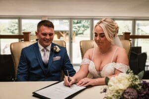 In a well-lit room, an elegant bride signs a document in her white gown and veil while her groom, dapper in a blue suit and tie, looks on. A bouquet accentuates the scene—captured beautifully through enchanting wedding photography that defines the charm of Moorlands Halifax.