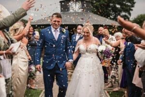 A bride and groom in formal attire walk hand in hand down an aisle, as captured by wedding photography. Guests on both sides shower them with confetti. A wooden structure with a clock stands in the background. The bride holds a bouquet of flowers, and both are smiling radiantly.