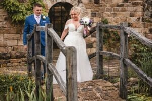 A bride in a white gown holding a bouquet and a groom in a blue suit walk across a small wooden bridge. Stone walls and greenery surround them, setting the perfect scene for moorlands wedding photography, capturing the rustic charm of Moorlands Halifax.