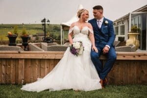 A bride in a white gown and a groom in a blue suit sit together on a wooden ledge, smiling at each other. Capturing this charming scene of wedding photography outdoors, the lush greenery of Moorlands Halifax provides the perfect backdrop as the bride holds her bouquet.