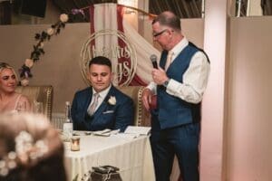 A man in a blue waistcoat and tie speaks into a microphone at the Moorlands Halifax wedding reception, standing beside a seated man in a suit. A decorative sign reads Mr & Mrs, surrounded by a white floral arrangement on the table—a perfect moment captured by wedding photography.