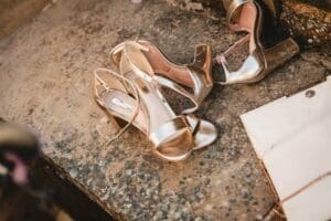 Two pairs of gold high-heeled sandals, perfect for wedding photography, rest on a textured stone surface. The sandals feature ankle straps and open toes, reflecting sunlight. Beige and pink tones of a nearby item are partially visible on the right.