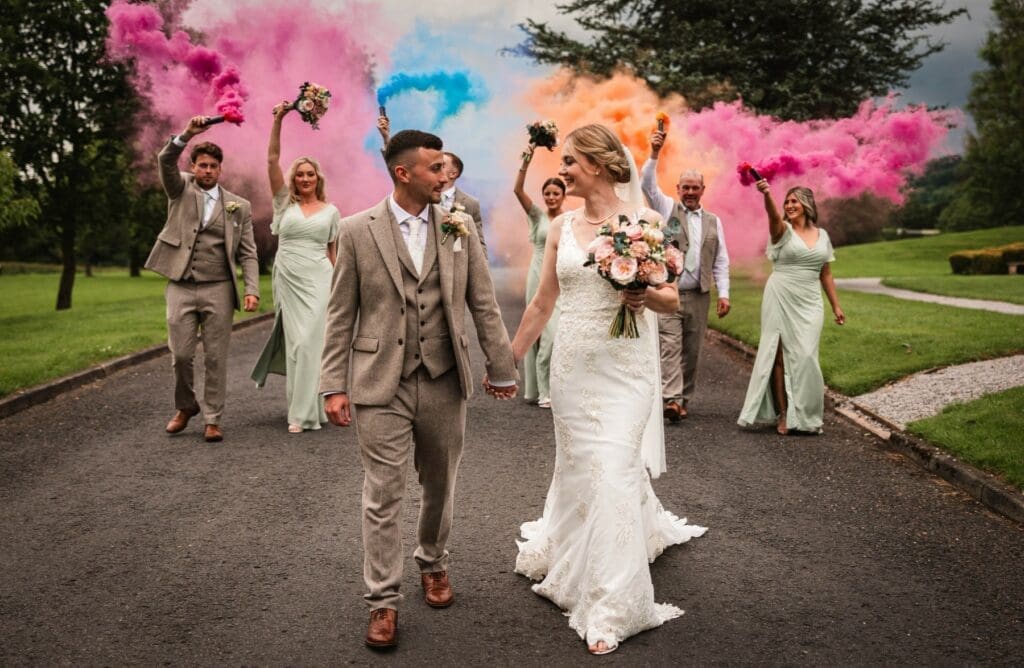 wedding party walking with smoke bombs behind in pink blue and orange, bridesmaids dressed in sage green outfits whilst the bride holds a beautiful pink bouqet