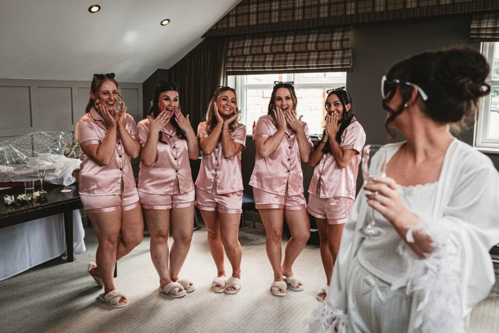 Five women in matching pink satin pajamas stand smiling and looking surprised, with hands on their faces. Another woman in a white robe, holding a glass, faces them in a bright, cozy room.
