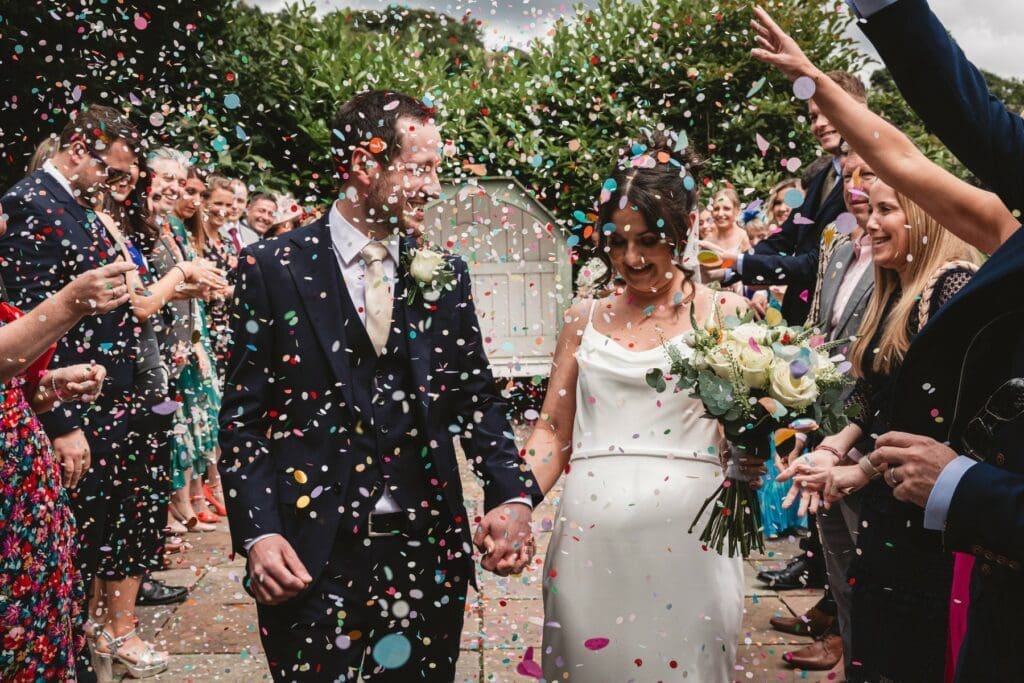 A newlywed couple walks hand in hand outdoors as guests shower them with the best wedding confetti. The bride holds a bouquet and smiles; the groom wears a dark suit. Friends and family line the path, celebrating joyfully.