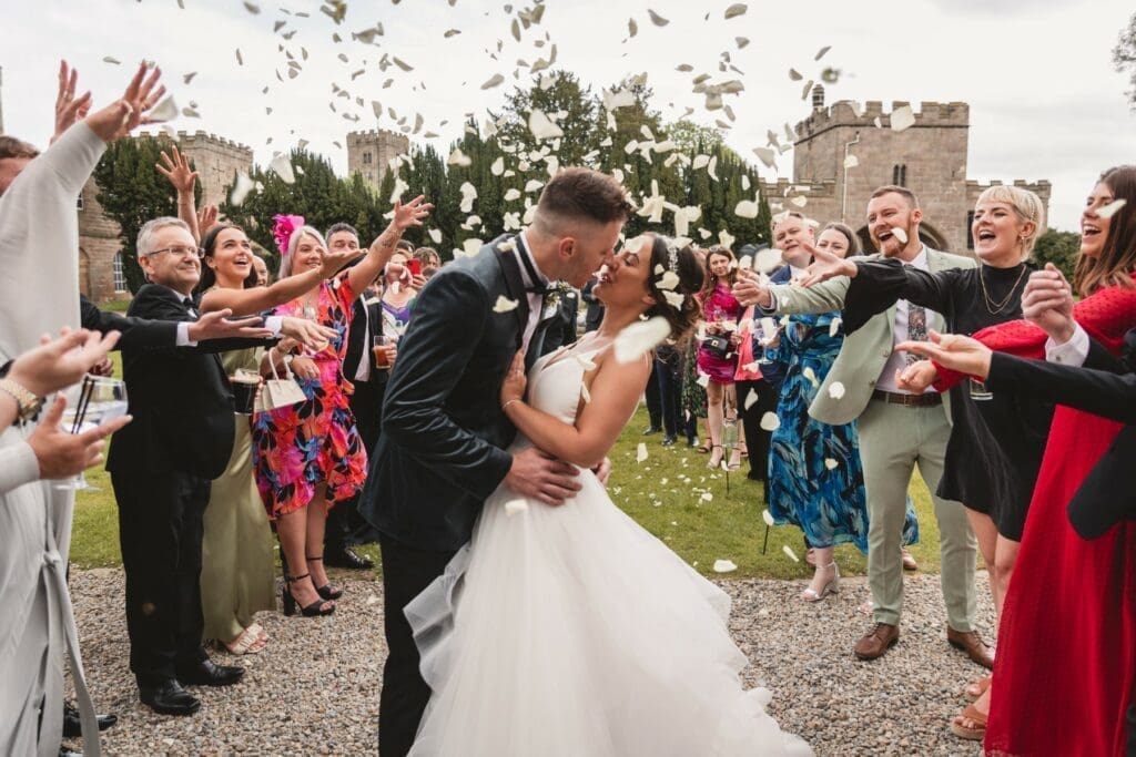 A bride and groom kiss outdoors as guests shower them with confetti and flower petals, celebrating amid joyful cheers and smiles in front of a stone castle—the best wedding moment.