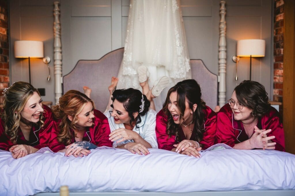 Five women in matching red robes, and one in white, lie on a bed laughing together. A white wedding dress hangs on the wall behind them, hinting at a joyful wedding preparation scene.