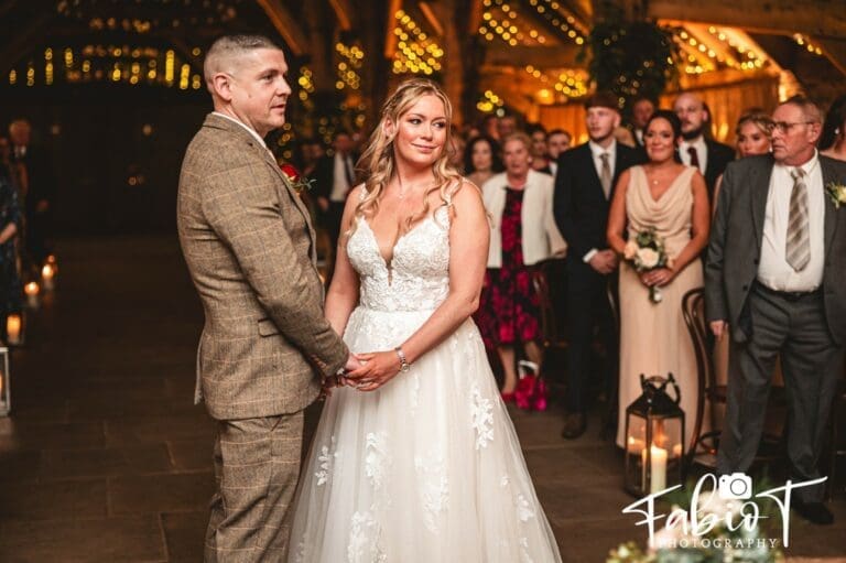 A bride and groom stand holding hands during their wedding ceremony in a warmly lit tithe barn venue, surrounded by formally dressed guests and wooden beams decorated with string lights.