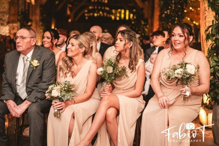 A group of people, including three women in matching light dresses holding bouquets and an older man in a suit, sit together smiling at a festive wedding in a tithe barn with warm lighting and floral decor.