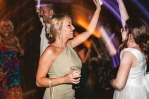 A woman in a light green dress smiles and dances with a drink in hand at a lively Tithe Barn Bolton Abbey party, surrounded by others celebrating under colorful lights.