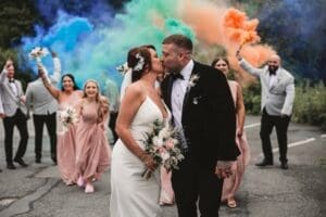 A bride and groom kiss outdoors, surrounded by their wedding party. Bridesmaids in pink dresses and groomsmen in light suits cheer, waving vibrant orange and blue smoke bombs—adding a dramatic touch to this joyful Smoke Bombs wedding celebration.