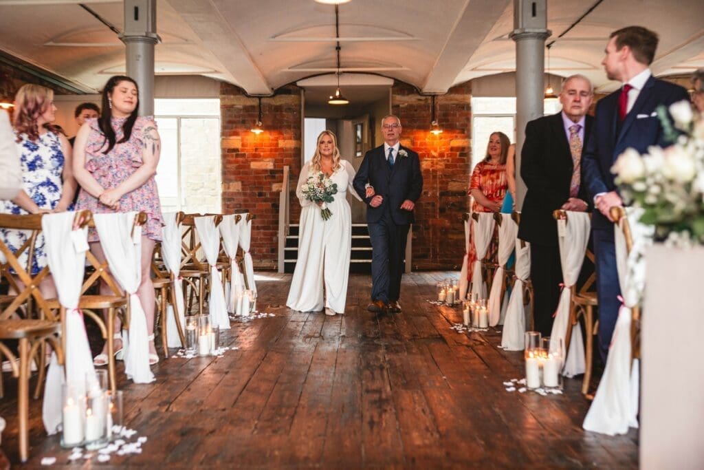 A bride walks down the aisle in the spinning room with her dad at the bowers mill