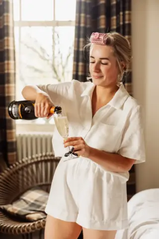 A woman in white pajamas with pink hair rollers smiles while pouring champagne into a glass at Holmes Mill, standing in a cozy room with plaid curtains and sunlight streaming through the window on her wedding morning.