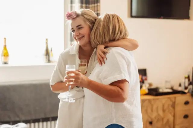 A woman wearing a hair roller and holding a glass of champagne smiles and hugs another woman in a bright room at Holmes Mill, Clitheroe, with bottles and sunlight in the background—capturing the joy of a wedding morning.