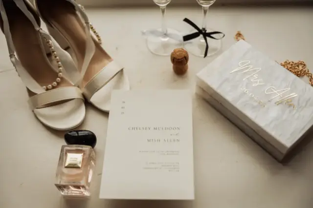 A flat lay of bridal accessories, including white heels with pearl straps, a wedding invitation, and a perfume bottle—perfect details for a Holmes Mill Clitheroe wedding—alongside two champagne glasses, a cork, and a white box labeled Mrs. Allen.