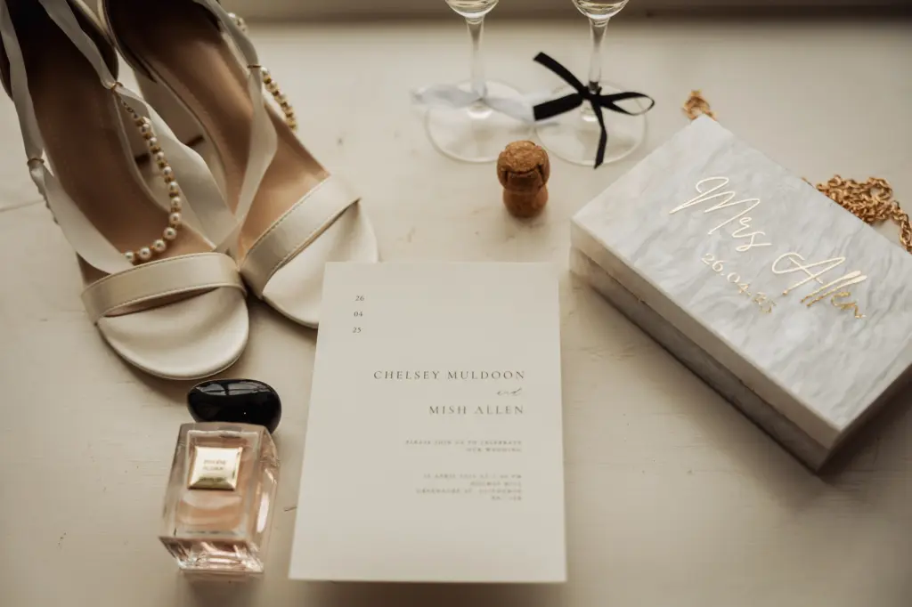 A flat lay of bridal accessories, including white heels with pearl straps, a wedding invitation, and a perfume bottle—perfect details for a Holmes Mill Clitheroe wedding—alongside two champagne glasses, a cork, and a white box labeled Mrs. Allen.