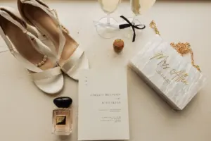 A flat lay of wedding accessories, including white high-heeled shoes, champagne glasses, a marble purse with Mrs. Allen in gold, perfume, and a wedding invitation for Chelsey Muldoon and Mish Allen at Holmes Mill in Clitheroe.