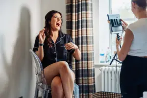 A woman in black pajamas laughs while sitting on a clear chair near a window at Holmes Mill, as another person stands nearby holding a ring light and phone, possibly taking a photo or video before the wedding in Clitheroe.
