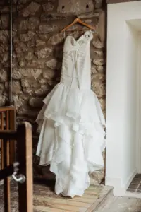 A white strapless wedding dress with a fitted bodice and layered ruffled skirt hangs on a wooden hanger against a rustic stone wall at Holmes Mill, Clitheroe, beside a white doorway.