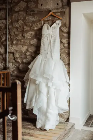 A white strapless wedding dress with a fitted bodice and layered ruffled skirt hangs on a wooden hanger against a rustic stone wall at Holmes Mill, Clitheroe, beside a white doorway.
