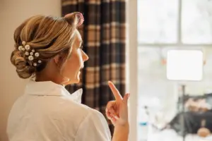 A woman with curled hair and pearl hairpins, wearing a white shirt, gestures while looking into a lit mirror near a window with plaid curtains at Holmes Mill in Clitheroe, preparing for a wedding.