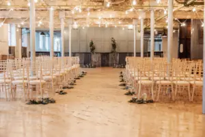 An indoor wedding ceremony at Holmes Mill, Clitheroe, features rows of light wooden chairs flanking a greenery-lined aisle under string lights in an industrial-style venue with exposed beams and columns.
